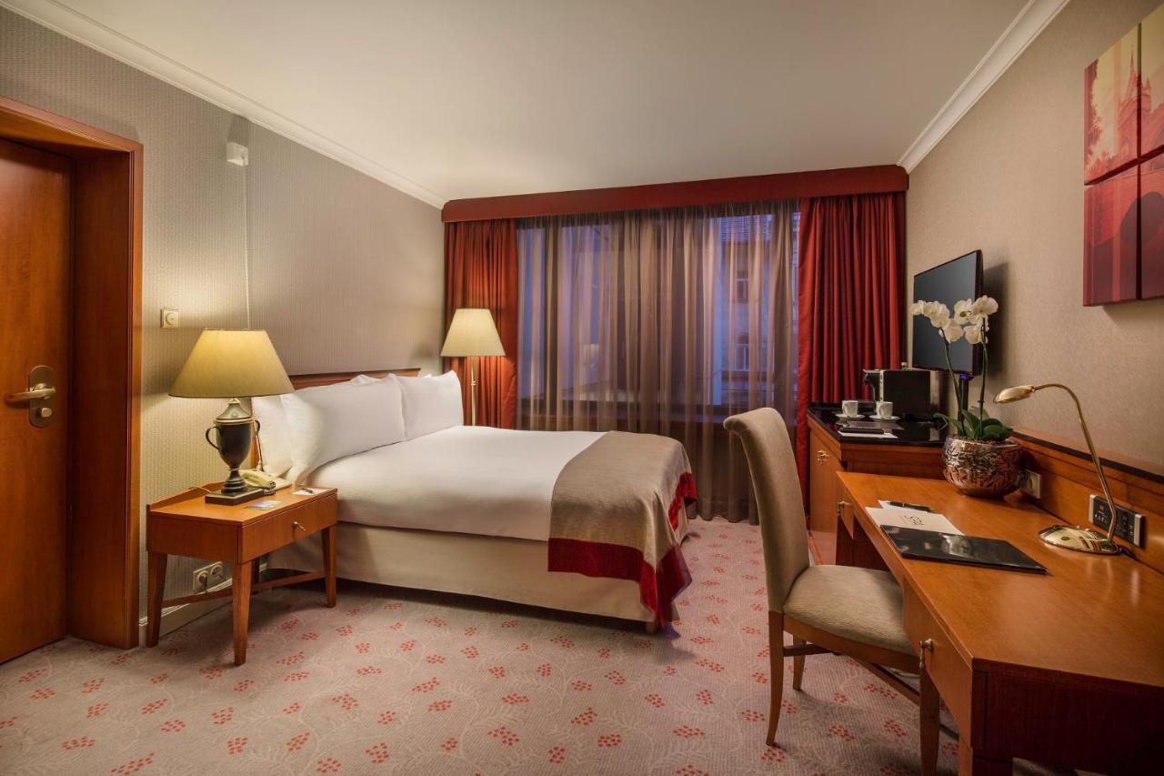 Intercontinental Budapest By Ihg Exterior foto The photo shows a well-furnished hotel room. It features a comfortable double bed with white linens and a decorative throw. There are two bedside tables with lamps on either side of the bed. A large window with sheer curtains lets in natural light, a