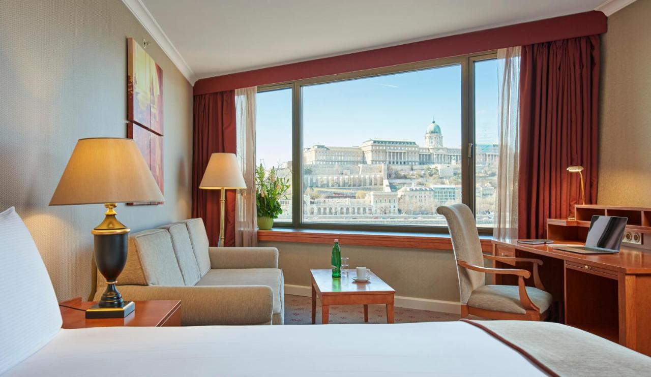 Intercontinental Budapest By Ihg Exterior foto The photo depicts a cozy hotel room featuring a large window with a scenic view. Inside, there are two comfortable chairs and a small table, with a lamp on a side table illuminating the space. A desk is also present, possibly for work. The window off