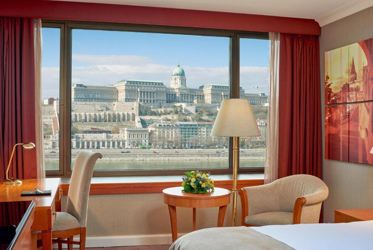 Intercontinental Budapest By Ihg Exterior foto The image shows the interior of a hotel room with a large window that offers a view of a picturesque landscape. Through the window, one can see a historic building situated on a hill, likely part of a castle or palace, and a scenic backdrop featuring