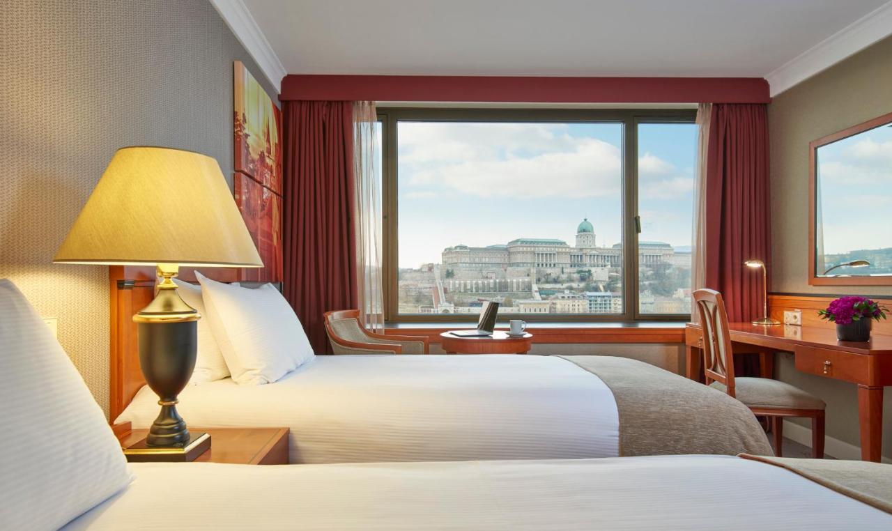 Intercontinental Budapest By Ihg Exterior foto The photo shows a hotel room featuring two beds with white linens and gray accents. There is a lamp on a bedside table, providing soft lighting. A large window offers a view of an outdoor scene, likely showcasing a cityscape with prominent buildings 