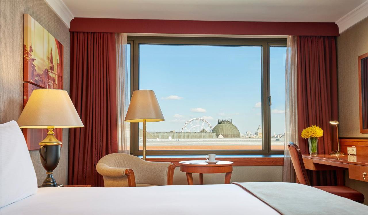 Intercontinental Budapest By Ihg Exterior foto The photo shows the interior of a hotel room with a view through a large window. In the foreground, there is a bed with white bedding, and nearby, a lamp and a small table with a chair. On the table, there's a cup, possibly for tea or coffee. The win