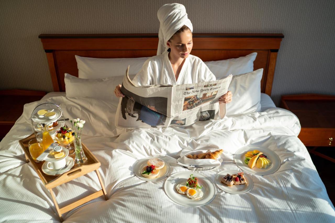 Intercontinental Budapest By Ihg Exterior foto The photo shows a person sitting on a bed, dressed in a white bathrobe with a towel on their head. They are reading a newspaper and surrounded by an assortment of food arranged on plates around them. There are various breakfast items, including pastr