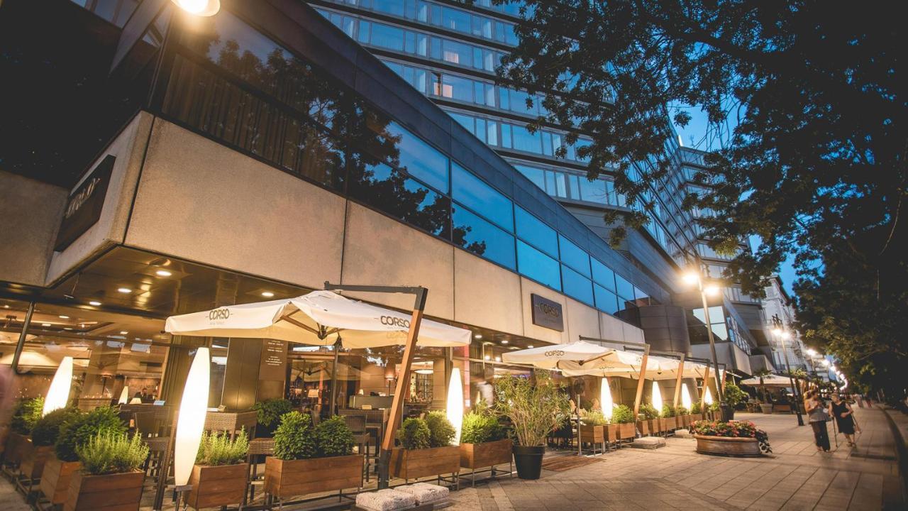 Intercontinental Budapest By Ihg Exterior foto The photo depicts a modern building with large glass windows, likely a restaurant or café, situated on a city street. It features an outdoor seating area with umbrellas providing shade, surrounded by plants and wooden planters. The scene is captured 