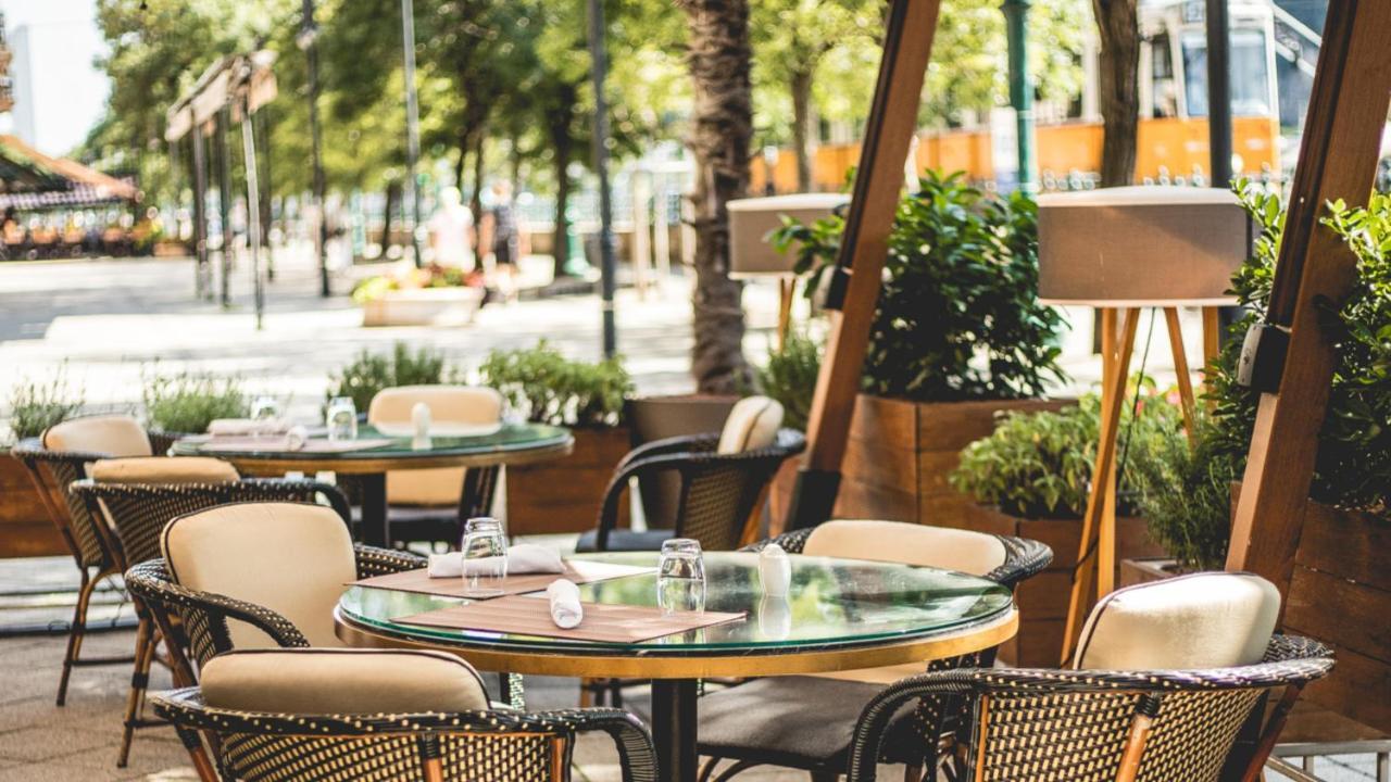 Intercontinental Budapest By Ihg Exterior foto The photo shows an outdoor dining area of a restaurant. There are several round tables set with chairs, some of which have cushions. The tables feature glass tops, and condiments or glassware can be seen on them. Surrounding the seating area are gree