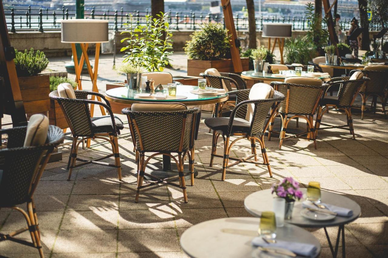 Intercontinental Budapest By Ihg Exterior foto The photo depicts an outdoor dining area featuring several round tables and chairs arranged on a patio. The tables are decorated with small flower arrangements and have place settings, suggesting a ready-to-dine setup. There are potted plants surroun