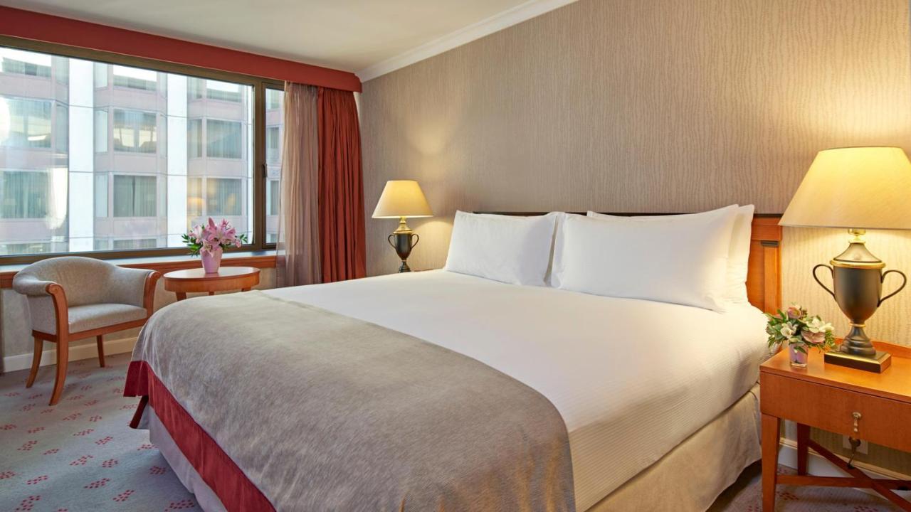 Intercontinental Budapest By Ihg Exterior foto The photo shows a hotel room with a large bed featuring white bedding and two pillows. The walls are light-colored with a subtle pattern, and there are large windows allowing natural light to enter. Next to the bed, there are bedside lamps providing 