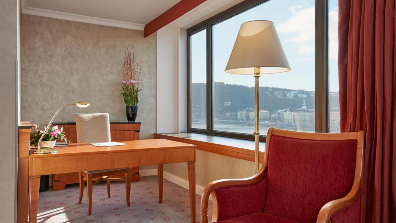 Intercontinental Budapest By Ihg Exterior foto The photo shows a stylish room, likely an office or a hotel suite. There's a wooden desk placed near a large window with a view of the outside landscape. A comfortable red armchair sits next to the desk, providing a cozy seating option. The room feat