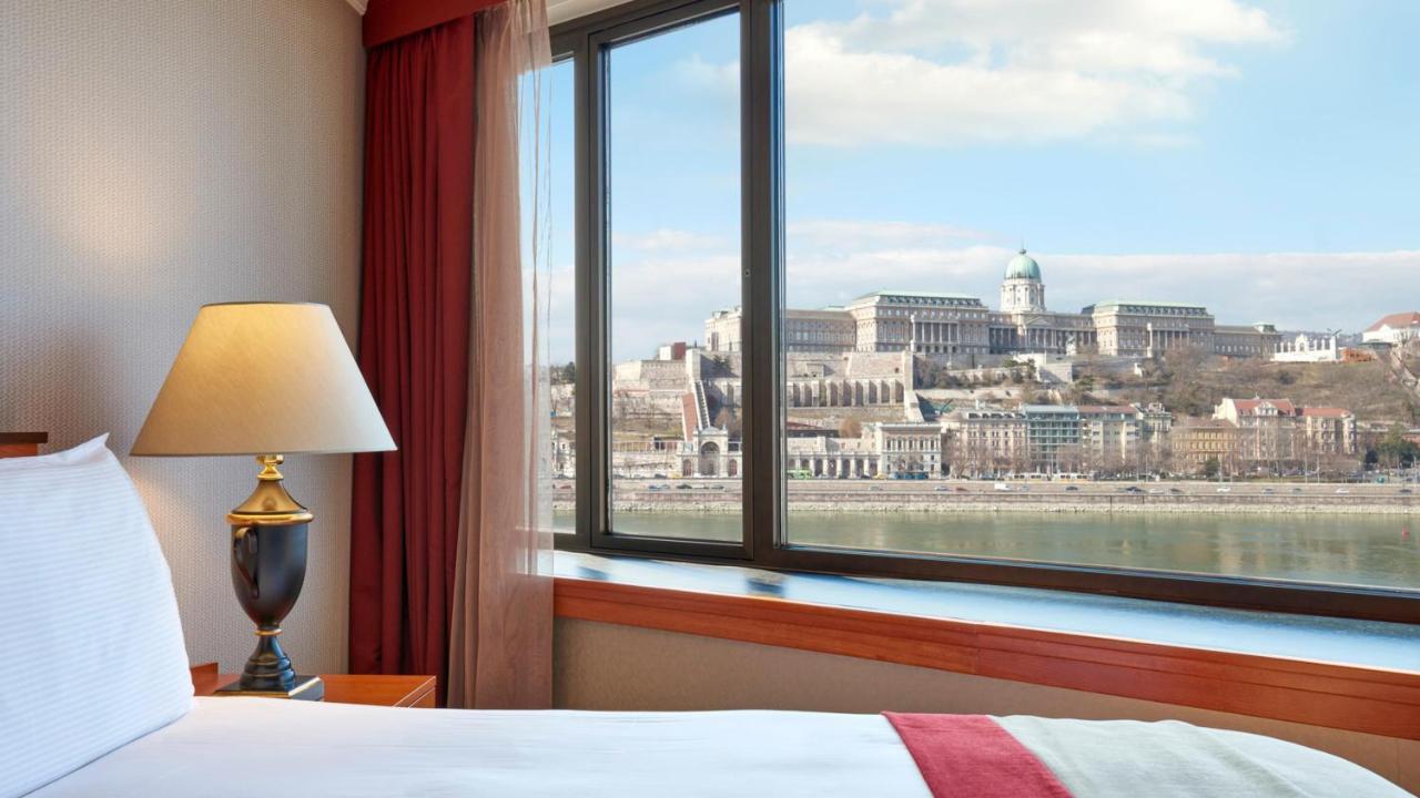 Intercontinental Budapest By Ihg Exterior foto The photo shows a view from a hotel room with a window overlooking a river and a historic building on a hillside. In the foreground, there is a bedside lamp, and you can see the soft furnishings in the room, such as a curtain and part of a bed. The l