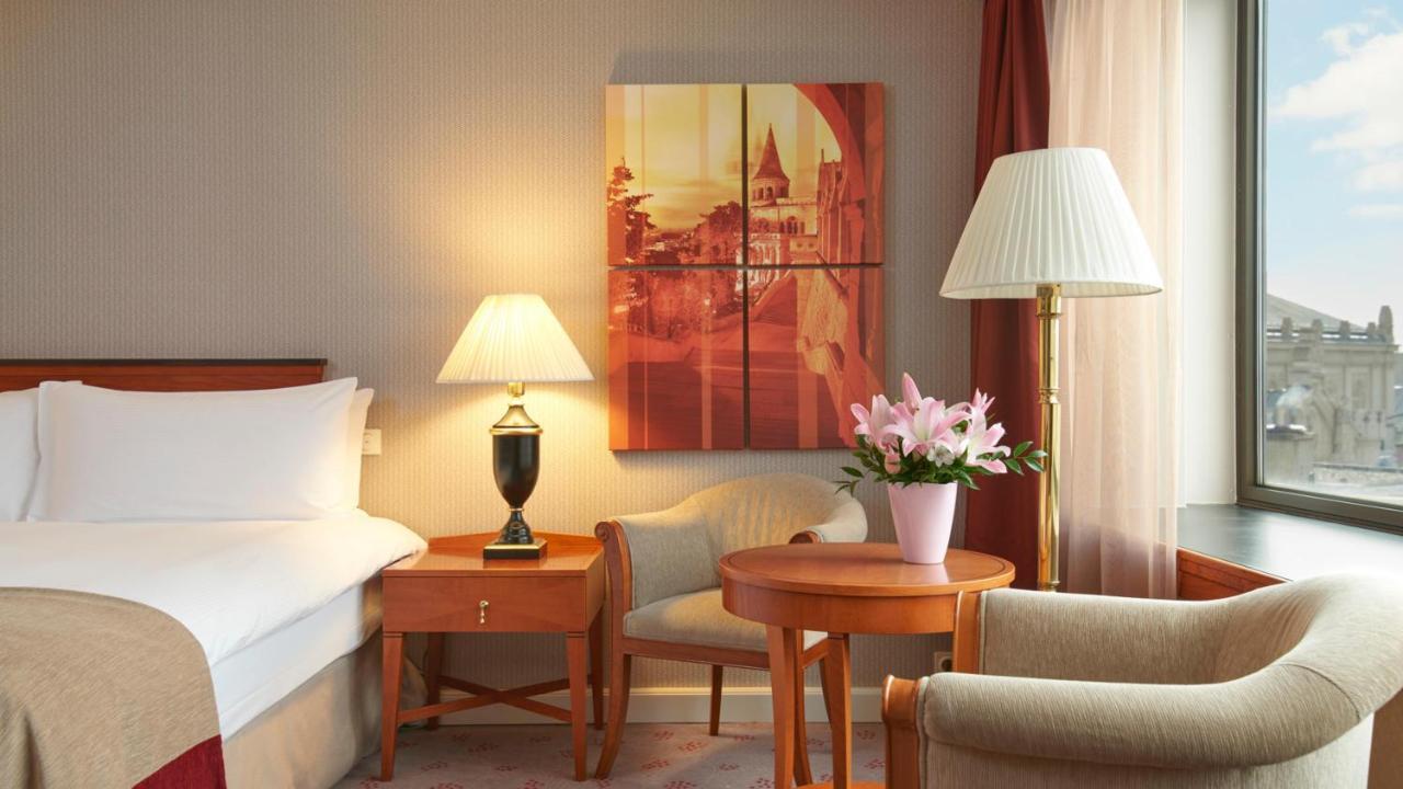 Intercontinental Budapest By Ihg Exterior foto The image shows a cozy hotel room featuring a bed with white linens, a small wooden table with a vase of pink flowers, and a comfortable chair. There's a decorative lamp on the table and another lamp next to a seating area. A piece of wall art depict