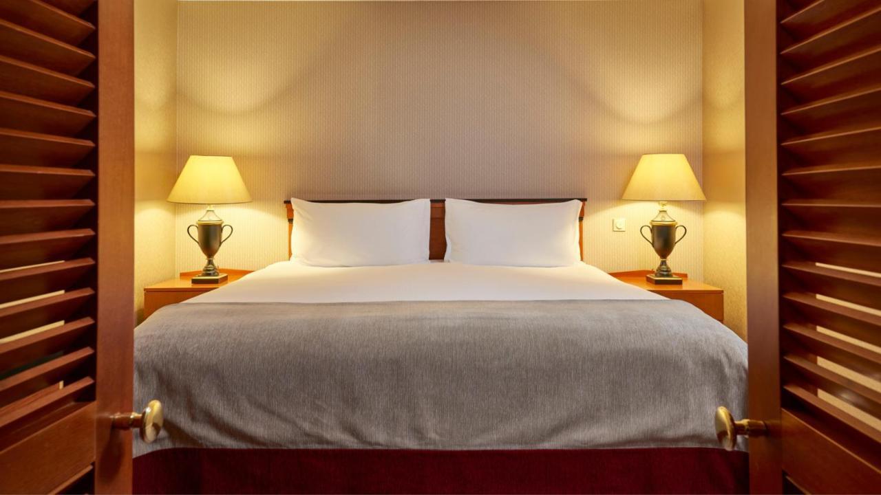 Intercontinental Budapest By Ihg Exterior foto The photo shows a cozy hotel bedroom with a large double bed centered in the frame. The bed has a neatly made white duvet and two white pillows. On either side of the bed, there are bedside tables with table lamps, adding a warm glow to the room. The