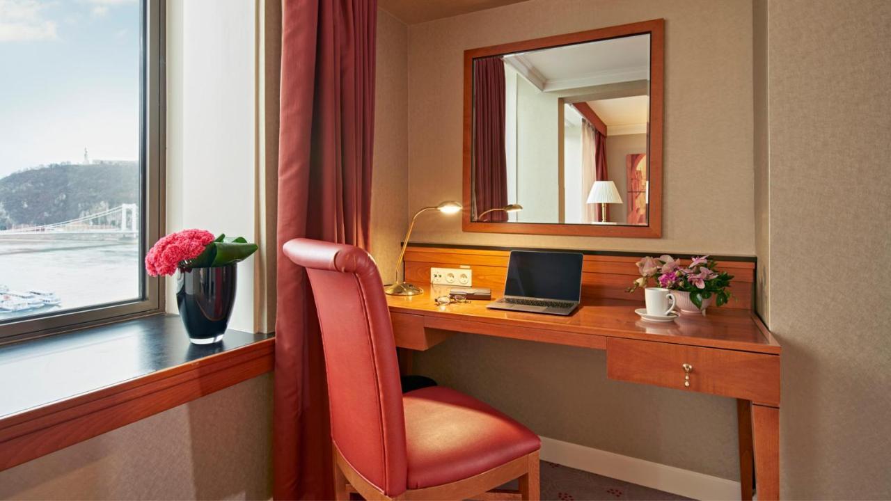 Intercontinental Budapest By Ihg Exterior foto The photo shows a small workspace set up in a room. It features a wooden desk with a laptop on it and a lamp for lighting. There is a comfortable red chair positioned at the desk. To the left of the desk, there is a window with a view, partially cove