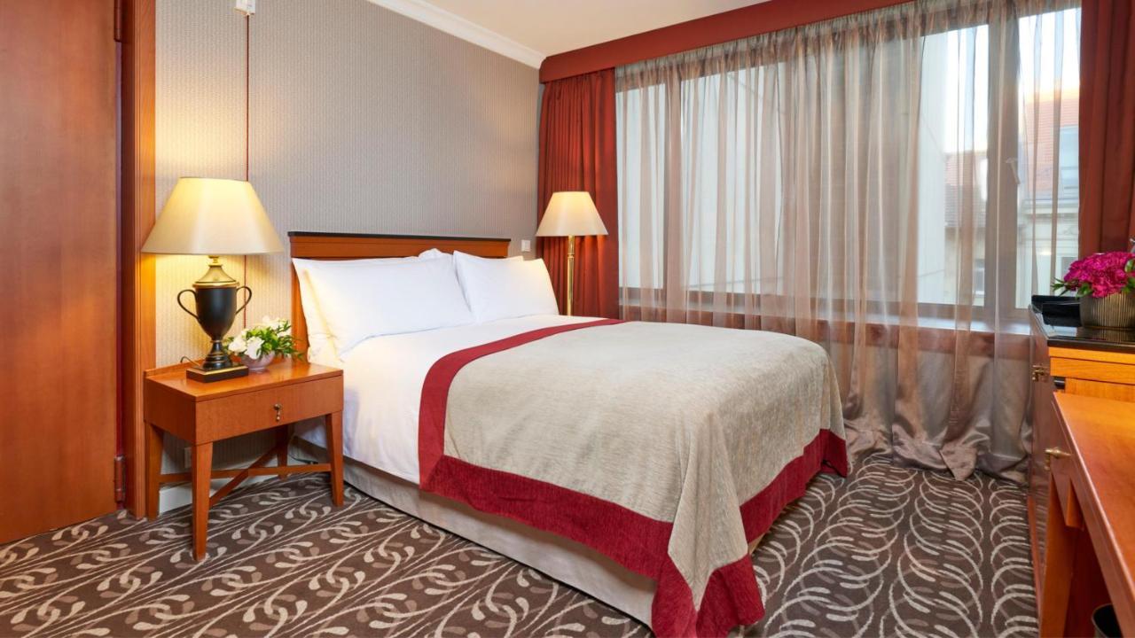 Intercontinental Budapest By Ihg Exterior foto The photo shows a hotel room featuring a neatly made bed with white linens and a light-colored blanket accented with a red border. There is a nightstand next to the bed with a decorative lamp and a small plant. A large window with sheer curtains allo