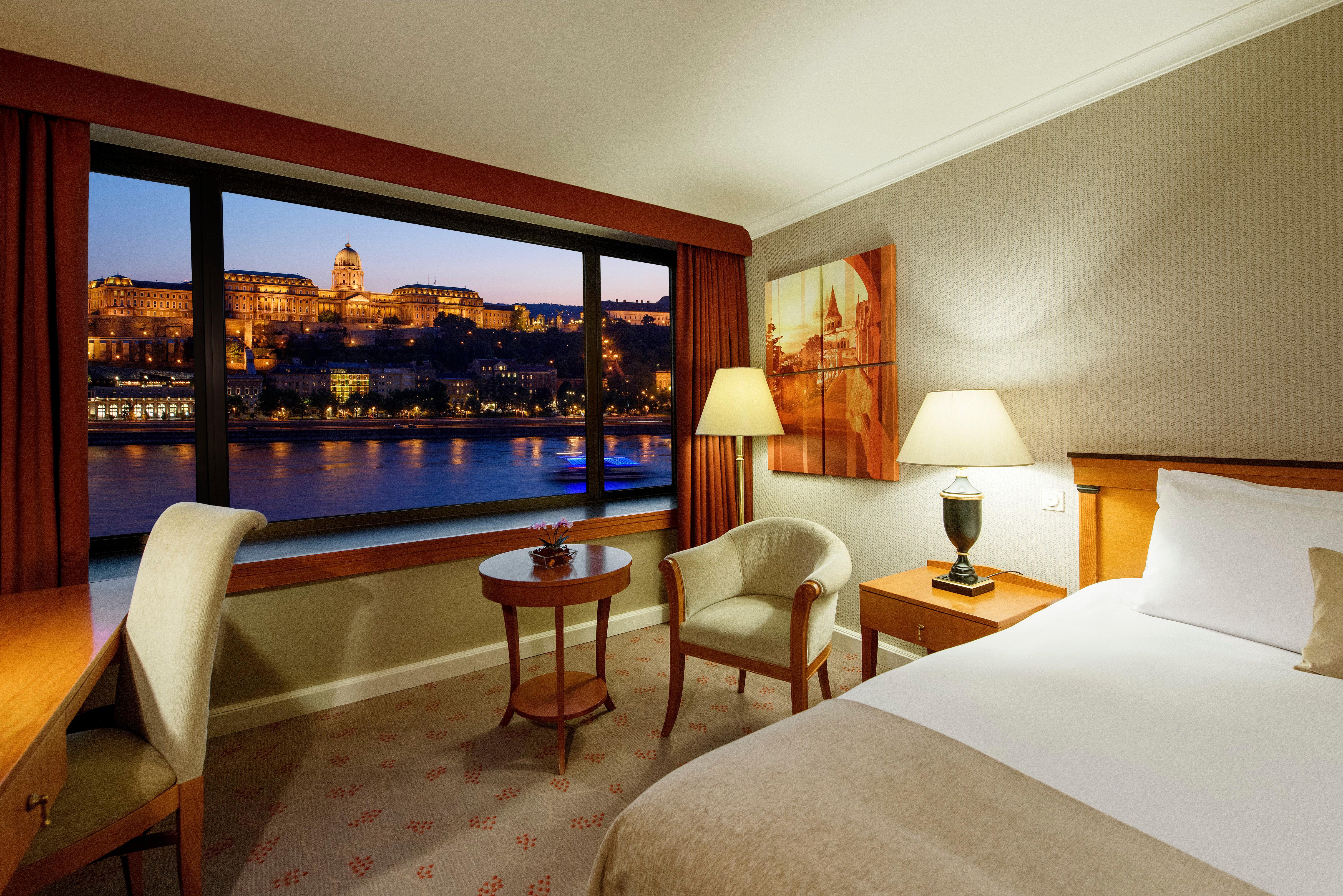 Intercontinental Budapest By Ihg Exterior foto The photo shows a hotel room featuring a cozy and well-lit interior. There is a comfortable bed with neutral-colored bedding positioned against one wall. To one side, there's a small wooden table with two chairs, creating a relaxing seating area. A s