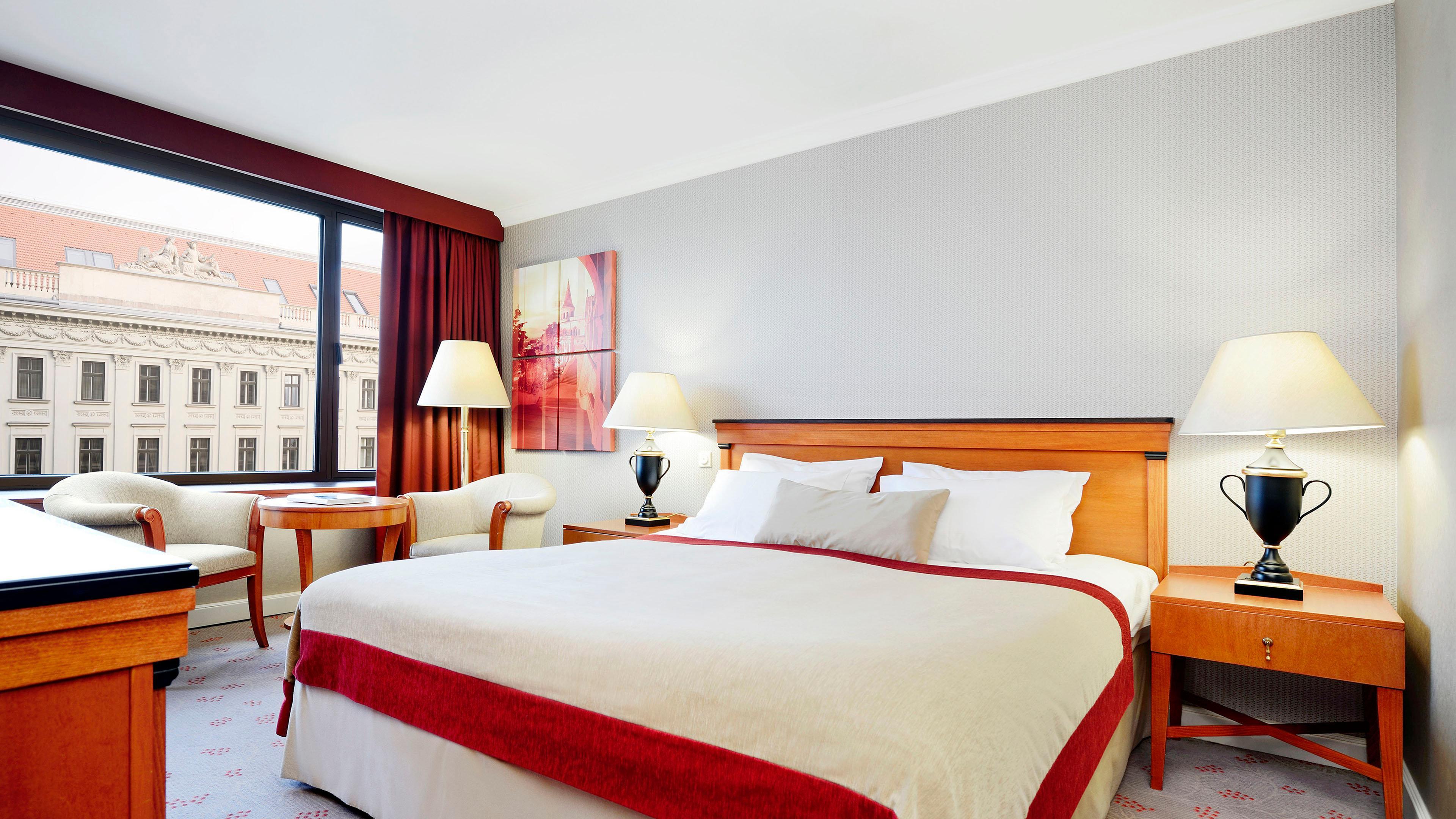 Intercontinental Budapest By Ihg Exterior foto The photo shows a hotel room featuring a large bed with white linens and a decorative red throw. There is a window with a red curtain, allowing natural light to enter the space. Next to the bed, there are two bedside tables with table lamps. A chair 