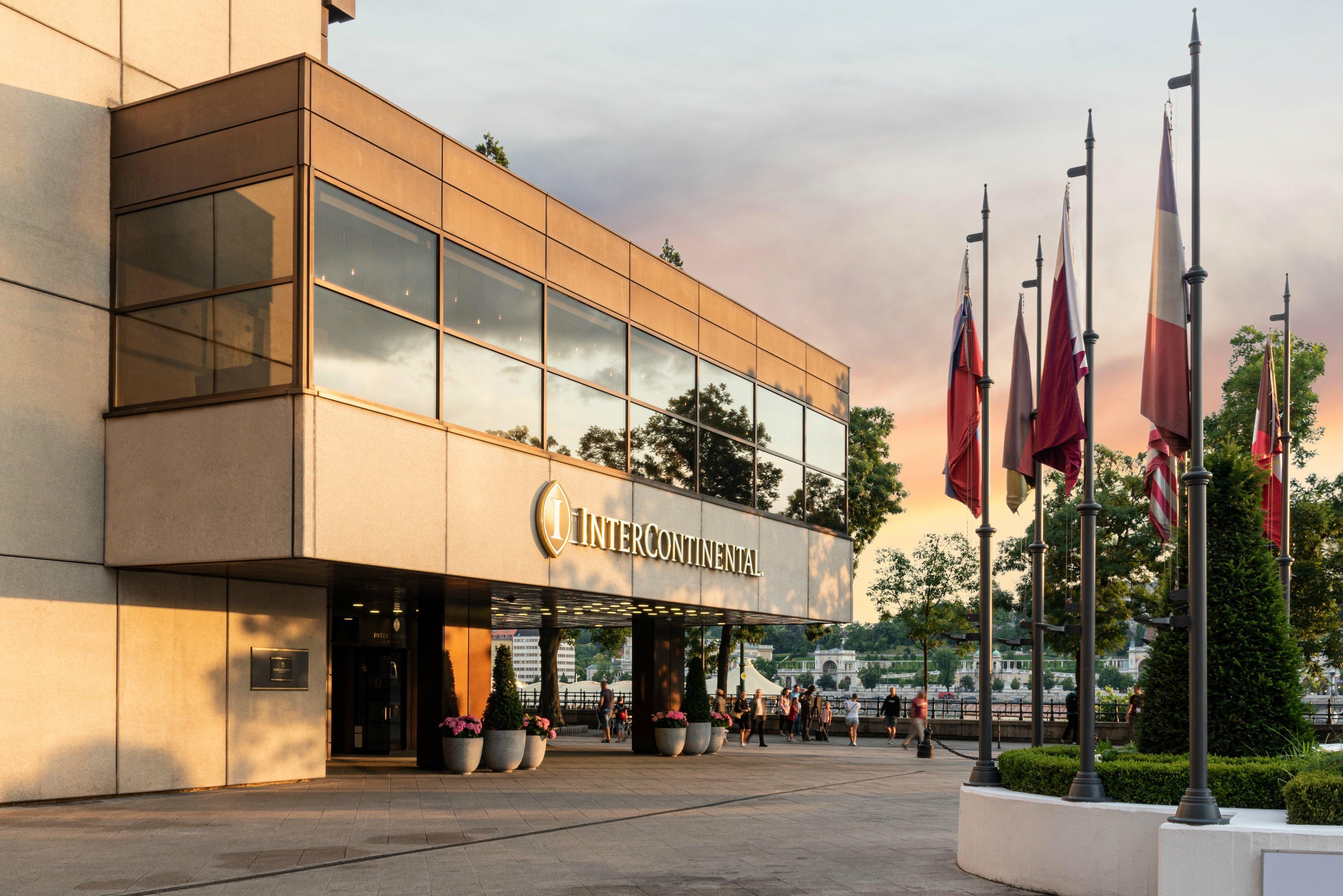 Intercontinental Budapest By Ihg Exterior foto The photo shows the exterior of a building, identified as the Intercontinental Hotel. It features a modern design with large glass windows and a warm color palette. In front of the building, there are several flagpoles displaying various flags. The s