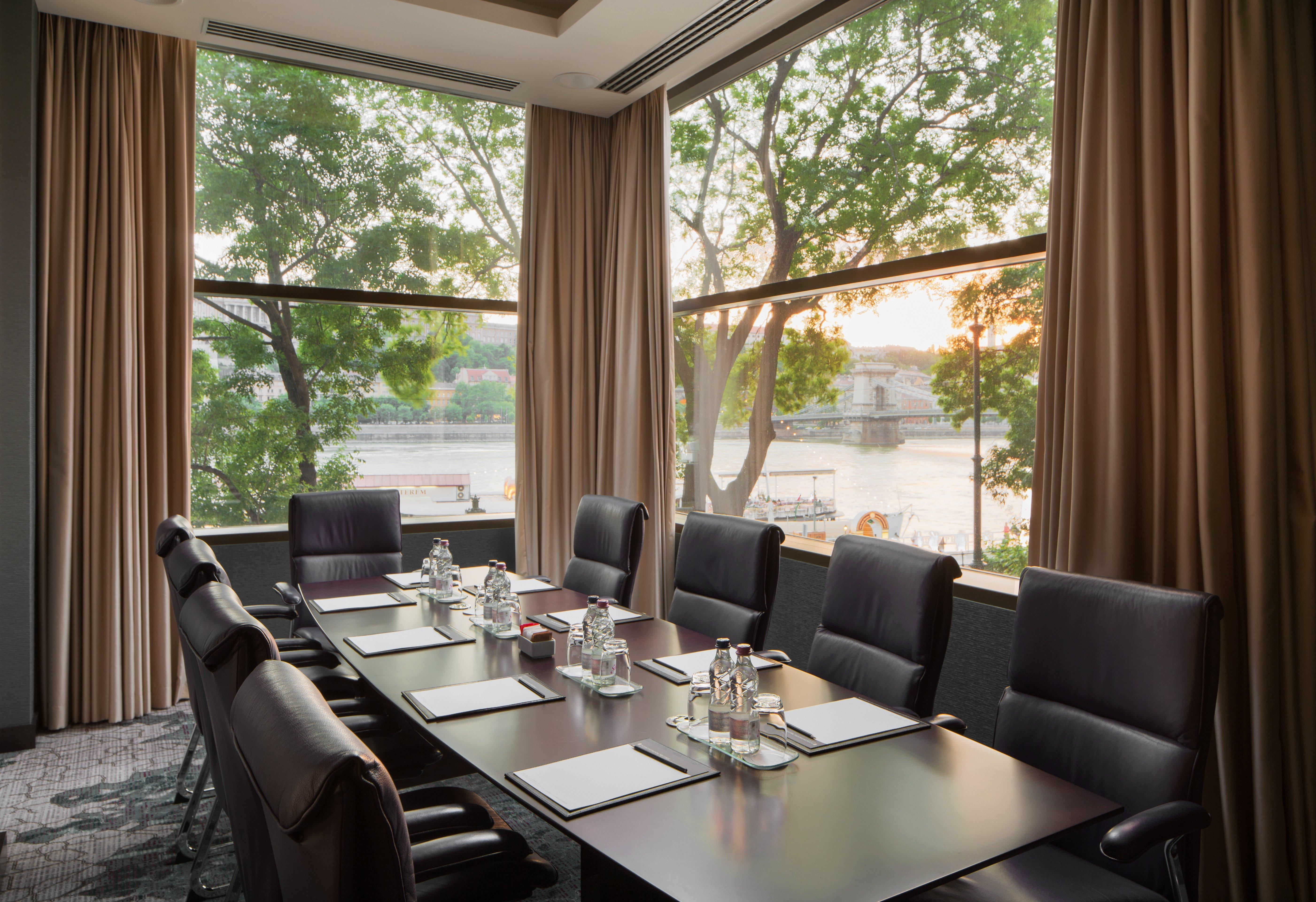 Intercontinental Budapest By Ihg Exterior foto The photo depicts a modern conference room with a long table surrounded by black office chairs. The table is set with glass water pitchers and drinking glasses. Large windows offer a view of greenery outside, with sunlight streaming in and casting a 