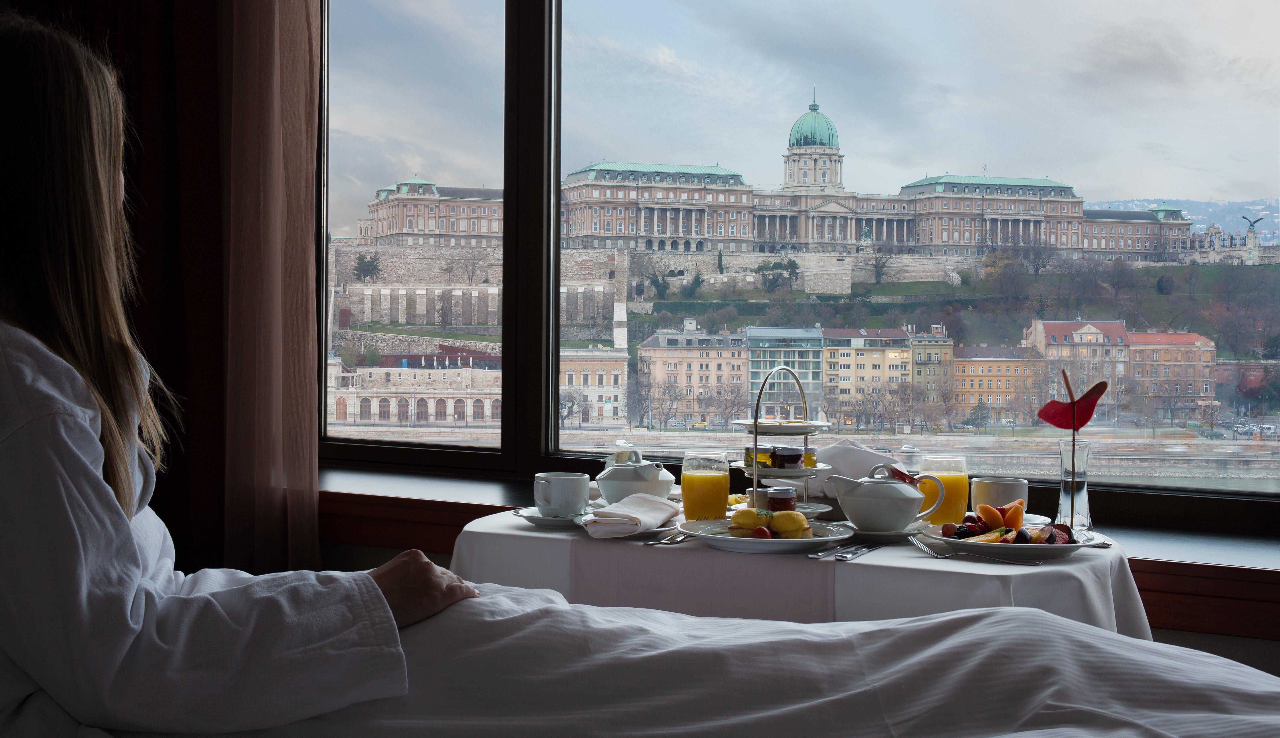Intercontinental Budapest By Ihg Exterior foto The photo depicts a serene breakfast scene in a hotel room, with a person in a bathrobe reclining on a bed. In the foreground, there is a neatly arranged breakfast spread on a table, including beverages and fruits, set against a backdrop of a beautif