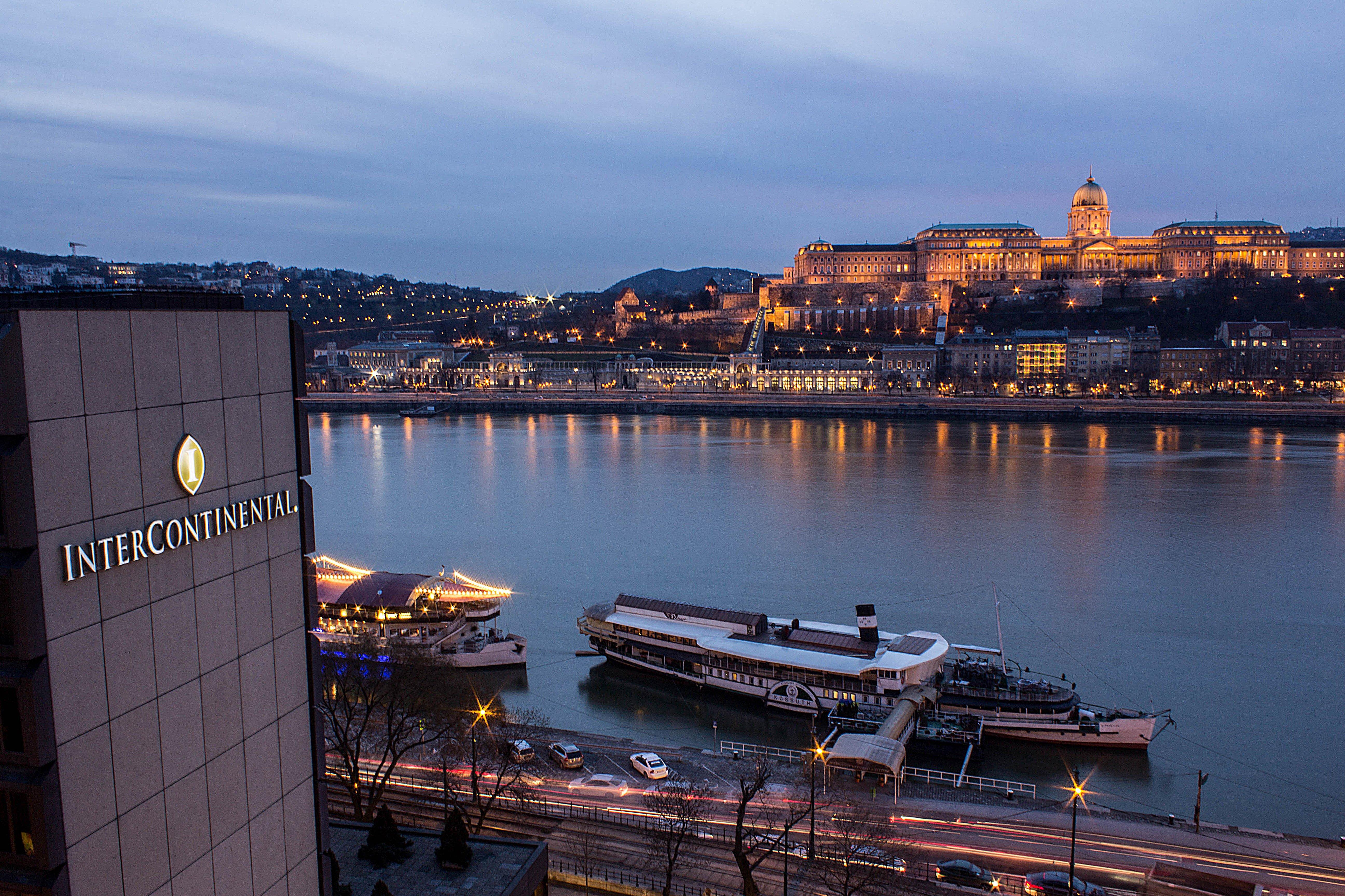 Intercontinental Budapest By Ihg Exterior foto The photo shows a nighttime view of a river, likely the Danube, with buildings illuminated along the shore. In the foreground, there is a hotel, identifiable by its logo, and in the background, a prominent structure atop a hill is lit up, possibly a 