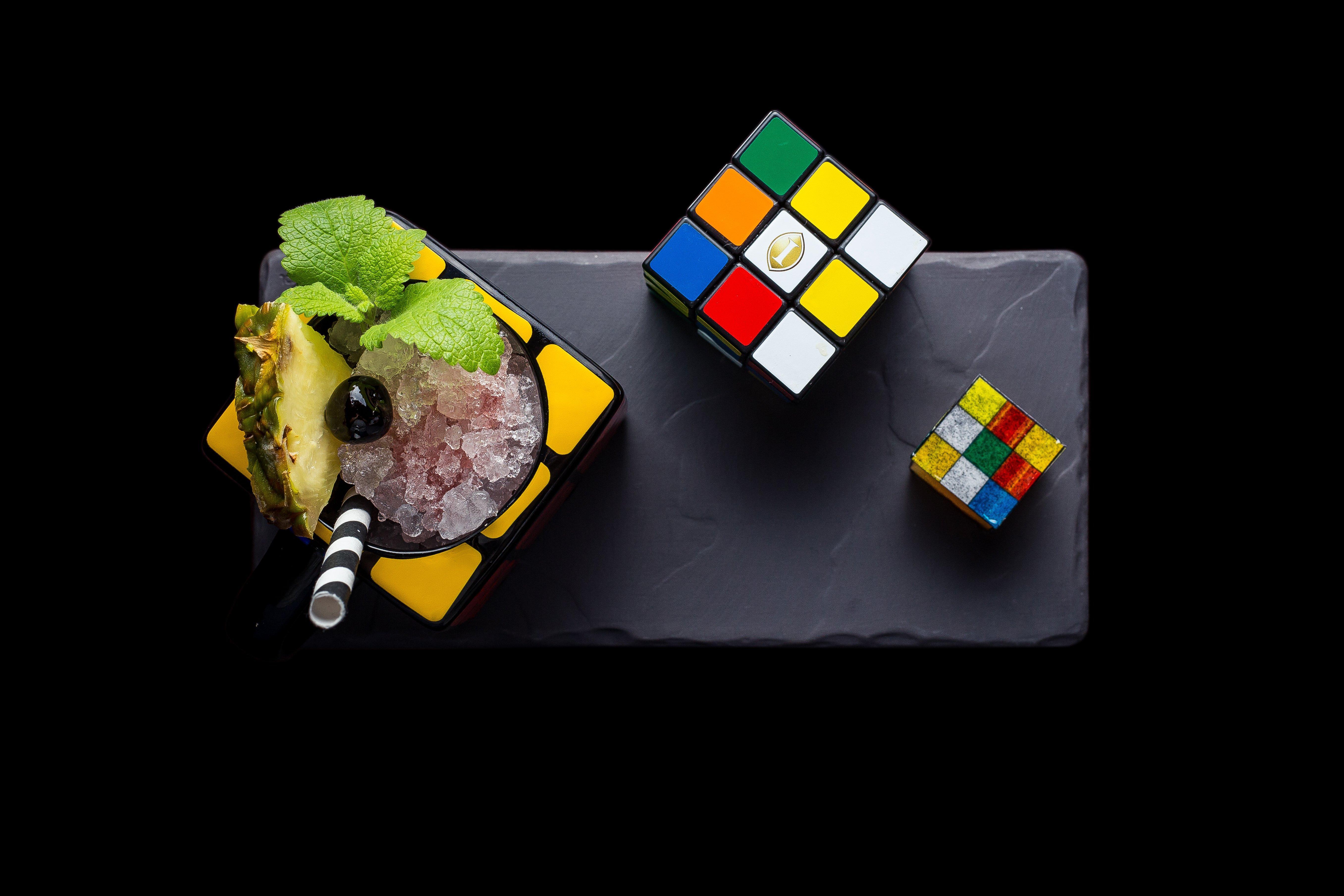Intercontinental Budapest By Ihg Exterior foto The photo shows a creative arrangement featuring a cocktail or dessert served in a colorful, square dish. The dish is garnished with mint leaves and a slice of pineapple, along with a black olive on top. Next to the dish, there are two Rubik's Cube p