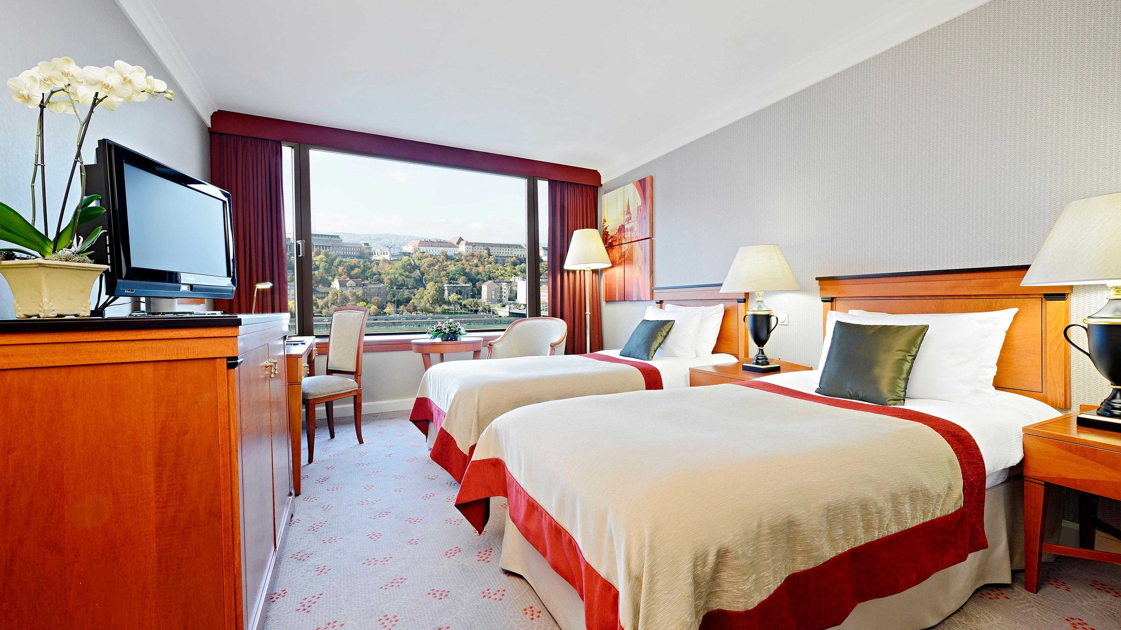 Intercontinental Budapest By Ihg Exterior foto The photo shows a hotel room featuring two twin beds with light-colored bedding and red accents. There is a large window allowing natural light to fill the space, with a view of an outdoor landscape. A wooden dresser and a television are positioned a