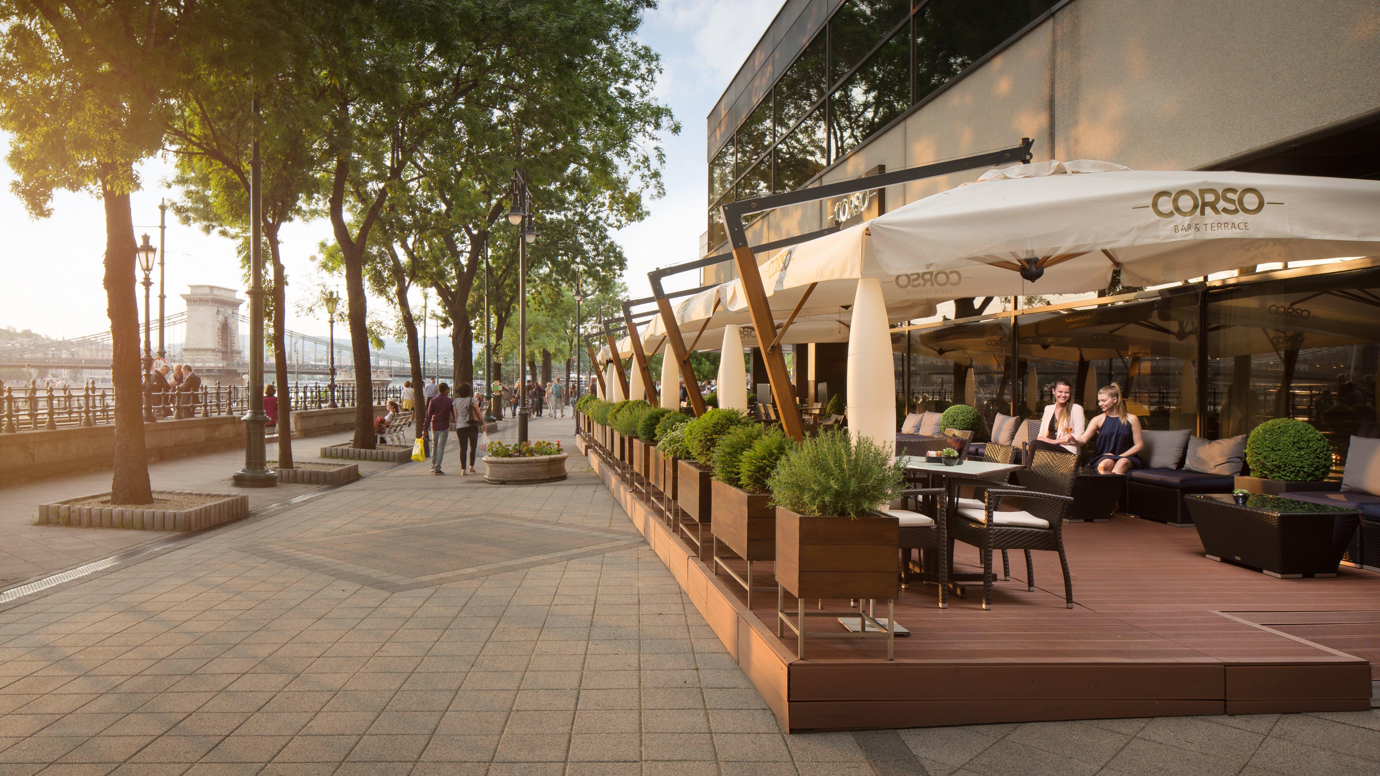 Intercontinental Budapest By Ihg Exterior foto The photo depicts an outdoor seating area of a café or restaurant. It features modern wooden furniture arranged under large, white umbrellas, providing shade for guests. Lush greenery and potted plants are visible along the seating area, enhancing th