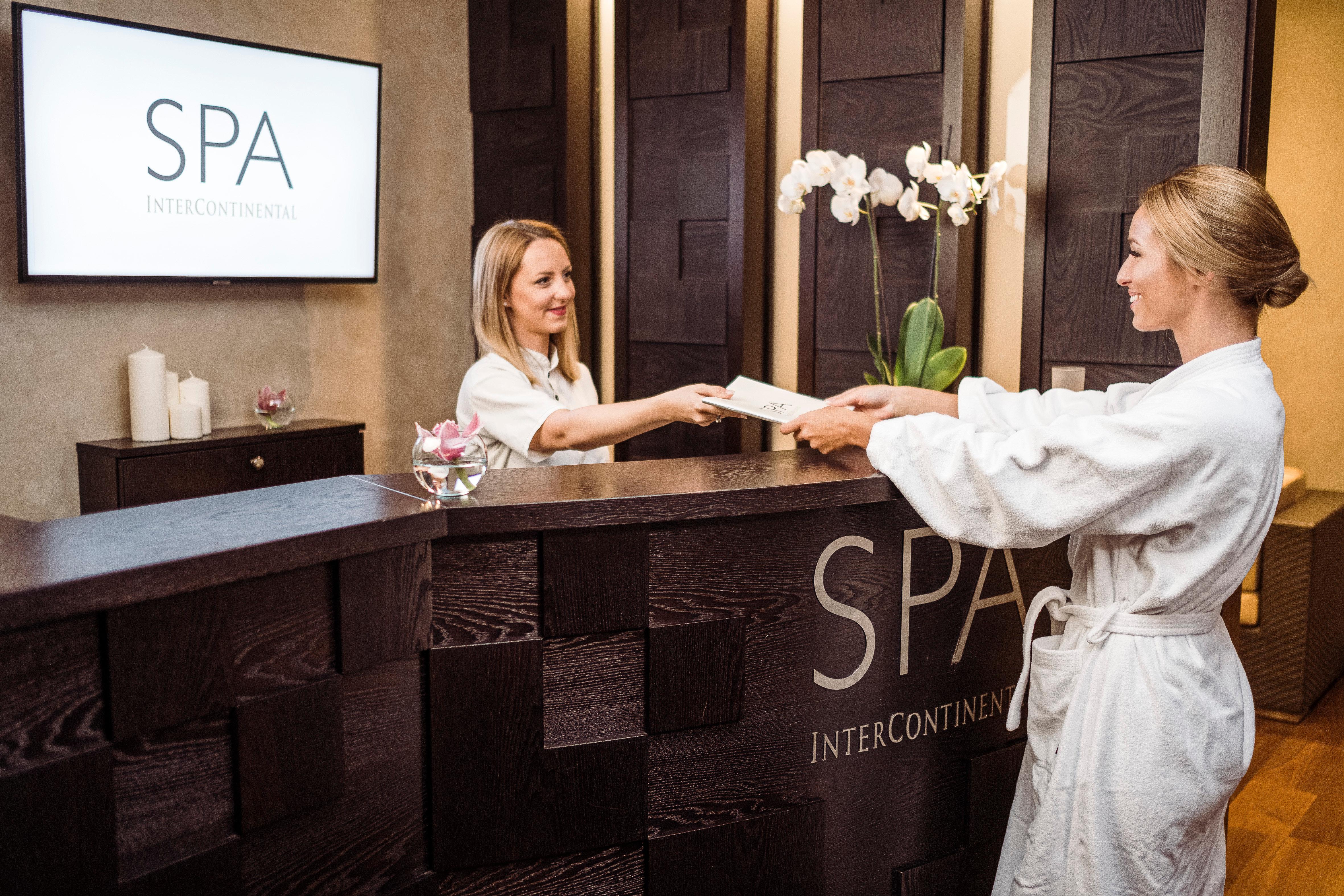 Intercontinental Budapest By Ihg Exterior foto The photo shows a spa reception area. A woman wearing a white robe is handing a document to a receptionist, who is also a woman. The receptionist is standing behind a stylish dark wooden desk that features the word "SPA" prominently displayed. The ba