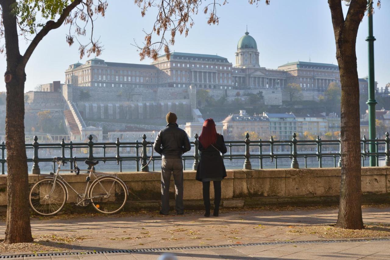 Intercontinental Budapest By Ihg Exterior foto The photo depicts two people standing by a pathway, overlooking a scenic view of a large building complex situated on a hillside, which likely represents a historic site or palace. The individuals are facing away from the camera, with one person wear