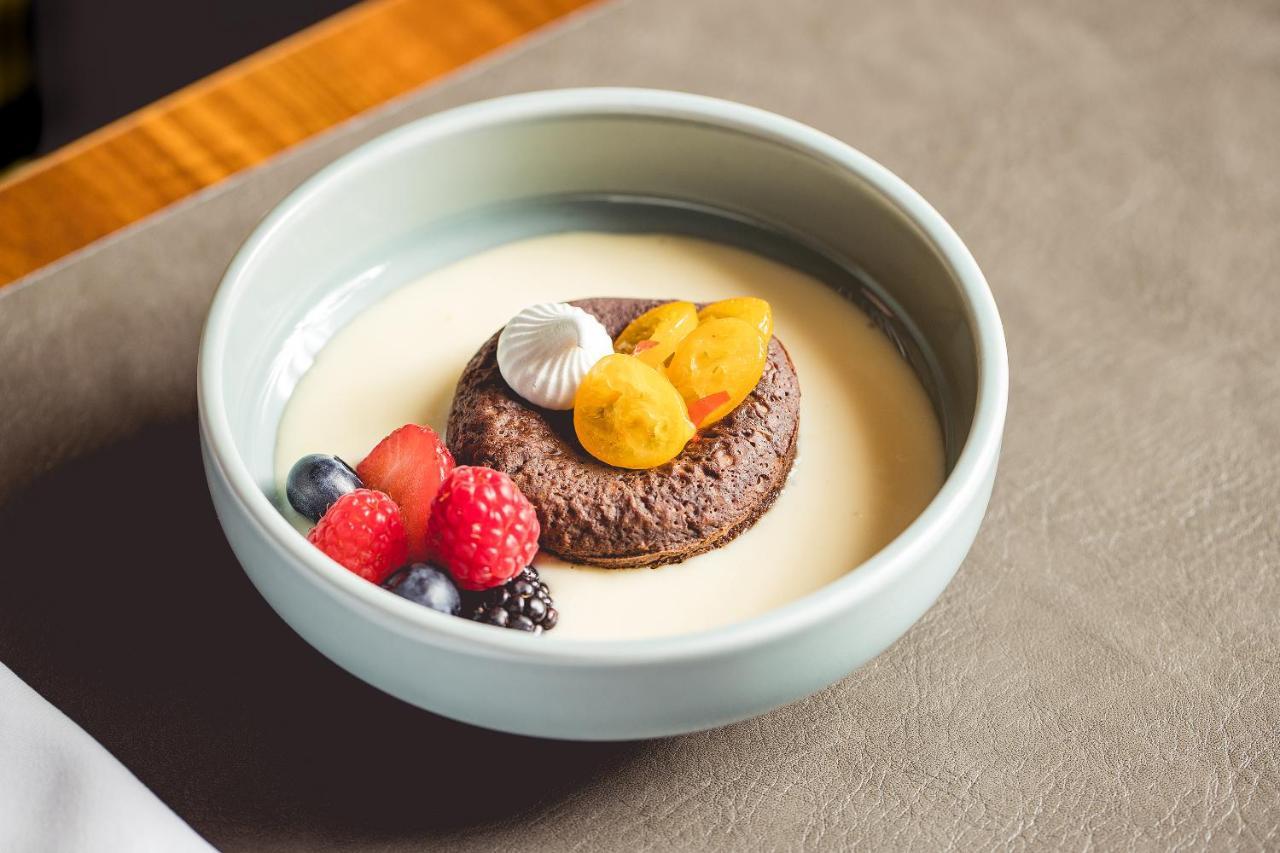 Intercontinental Budapest By Ihg Exterior foto The photo shows a dessert presented in a light blue bowl. At the center, there is a round chocolate cake or cookie topped with small pieces of yellow fruit, possibly apricots or peaches. Surrounding the cake are various fresh berries, including raspb